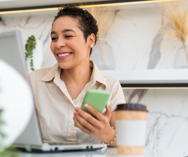 Smiling woman working at laptop and holding phone with a to-go coffee cup